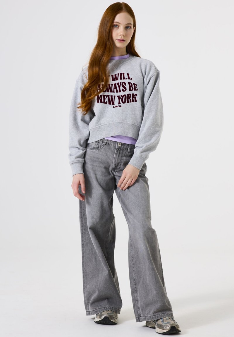 Sudadera gris corta con texto burdeos "SIEMPRE SERÉ NUEVA YORK" sobre una camiseta de manga larga lavanda, combinada con jeans grises de pierna ancha.