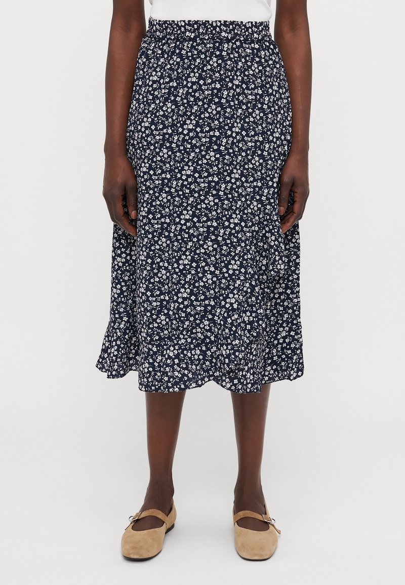 Femme portant une jupe midi bleu marine à imprimé floral blanc, associée à des chaussures plates Mary Jane beige, debout devant un fond uni.