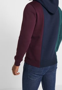 Sudadera con capucha que presenta un diseño a bloques de color con secciones de burdeos, azul marino oscuro y verde. Fabricada con un tejido suave y texturizado, con puños acanalados.