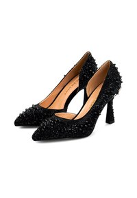 Zapatos de tacón alto negros con punta afilada, adornados con picos y strass sobre una superficie texturizada. Presentan un tacón delgado y lados abiertos.