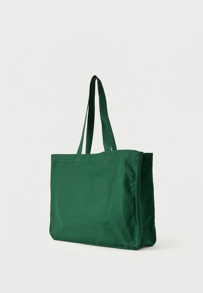 Πράσινη τσάντα tote από ύφασμα με δύο χειρολαβές και μια μπροστινή τσέπη, στέκεται σε απλό λευκό υπόβαθρο.