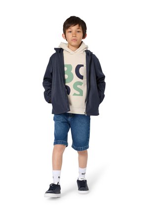 Marineblaue Jacke mit Reißverschluss aus strukturiertem Stoff, beige Kapuzenjacke mit grünem und marineblauem Muster, Jeansshorts, weiße Socken mit Logo und dunkle Sneakers.