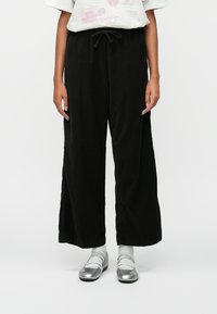 Fekete, széles lábú nadrág texturált anyagból, húzózsinórral a derék részen. Ezüst cipőkkel és szürke zoknikkal párosítva. Hétköznapi öltözet bemutatása.