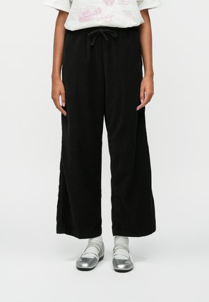 Pantaloni negri cu croi larg, dintr-un material texturat, cu talie ajustabilă prin șnur. Asortați cu pantofi argintii și ciorapi gri. Prezentare a unei ținute casual.