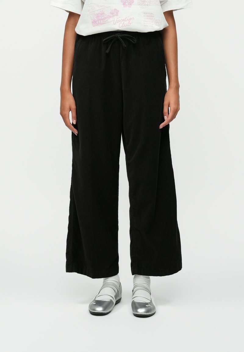 Fekete, széles lábú nadrág texturált anyagból, húzózsinórral a derék részen. Ezüst cipőkkel és szürke zoknikkal párosítva. Hétköznapi öltözet bemutatása.