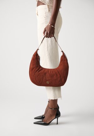 Suede brown shoulder bag with a curved design, gold hardware, and a smooth texture; paired with white cropped pants and black stilettos.