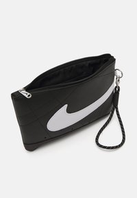 Crna sintetička torbica za rukovanje s sivim Nike logotipom. Ima zatvaranje na zatvarač i uklonjivi remen za zapešće ukrašen bijelim točkicama. Tekstura površine.