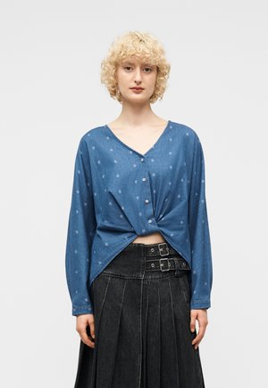 Femme aux cheveux blonds bouclés portant un chemisier bleu à boutons avec de petits motifs et un devant torsadé, assorti à une jupe plissée noire ceinturée.