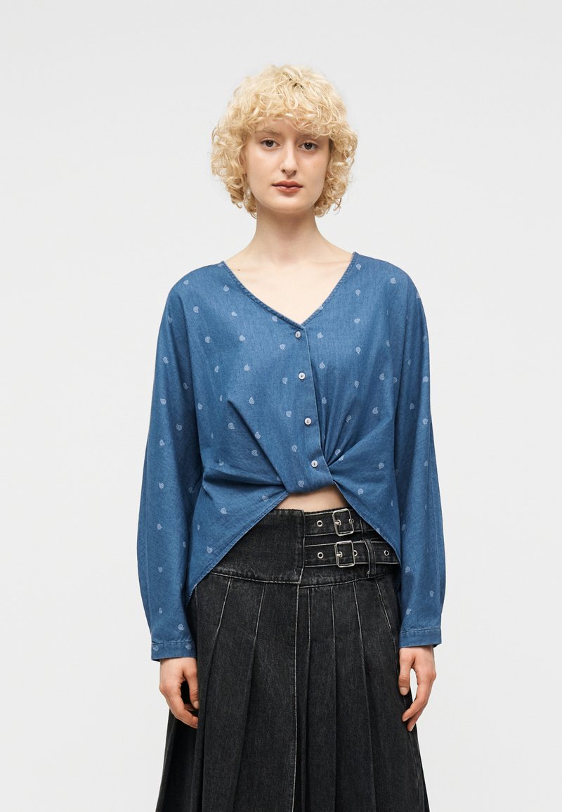 Γυναίκα με σγουρά ξανθά μαλλιά που φοράει μπλε πουκάμισο με κουμπιά και μικρά σχέδια, με στριφογυριστό μπροστινό μέρος, συνδυασμένο με μαύρη πλισέ φούστα με ζώνη.