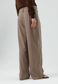 Wijde broekspijpen in een lichtbruine stof, met een gladde textuur, een op maat gemaakte pasvorm en zijzakken. De tailleband heeft een knoopsluiting.