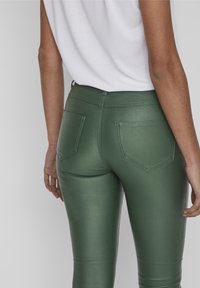 Pantalones verde de cintura alta hechos de un material brillante y elástico. Presentan dos bolsillos traseros con detalles de costura. Textura suave y diseño ajustado.