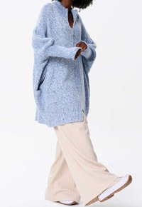 Ljusblå oversize stickad cardigan med hög krage, dragkedja framtill, ribbade muddar och sidofickor, bärs över beige byxor med smala ränder.