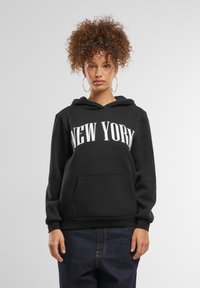 Fekete kapucnis felső, melyen fehér "NEW YORK" felirat található, kanguru zsebbel, húzózsinórral ellátott kapucnival és puha textúrával. Kék farmernadrággal párosítva.
