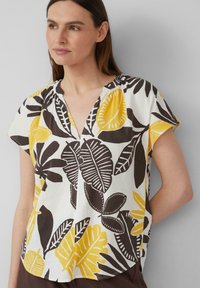 Blouse à manches courtes en tissu blanc avec des motifs de feuilles noirs et jaunes. Design à col en V avec une coupe décontractée et un ourlet arrondi.