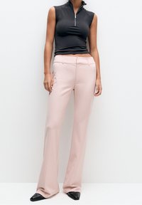 Haut noir sans manches avec col et fermeture éclair, associé à un pantalon évasé rose pâle. La tenue présente une texture lisse et une silhouette ajustée.