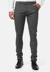 Pantalon gris sur mesure en tissu texturé, avec une coupe slim, des poches avant et une fermeture à boutons. Porté avec des chaussures noires.