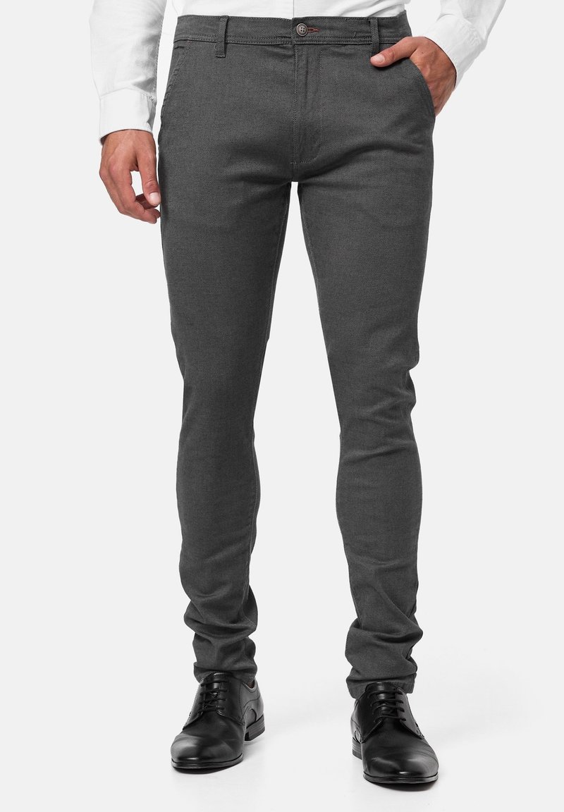 Pantalon gris sur mesure en tissu texturé, avec une coupe slim, des poches avant et une fermeture à boutons. Porté avec des chaussures noires.