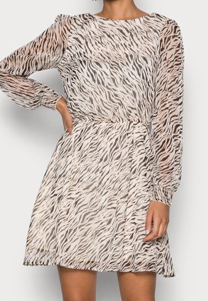 Donna con vestito corto trasparente a maniche lunghe con stampa zebra beige e nera, vita e polsini elastici, mano sul fianco.