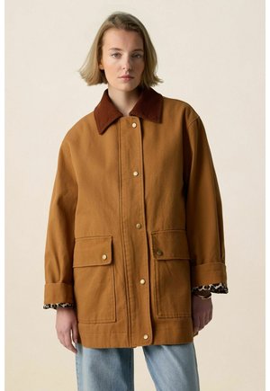 Femme portant un manteau marron boutonné avec de grandes poches avant et un col sombre, debout devant un fond clair uni.