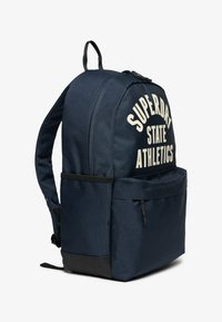 Sac à dos bleu marine avec le texte imprimé blanc "SUPERDRY STATE ATHLETICS." Comprend une poche zippée à l'avant et un panneau d'accent noir en bas.