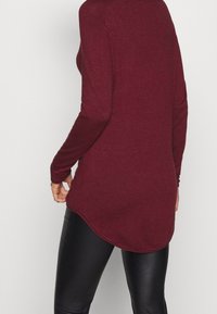 Maglia bordeaux a maniche lunghe con orlo curvo, realizzata in tessuto morbido, con maniche attillate e una vestibilità rilassata, abbinata a pantaloni neri.