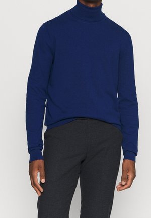 Homme portant un pull à col roulé bleu marine ajusté et un pantalon à carreaux gris foncé, debout devant un fond uni.
