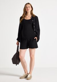 Top nero aderente abbinato a una camicia nera con bottoni, pantaloni corti abbinati, scarpe beige e una borsa marrone con texture. Tessuto liscia, design casual.