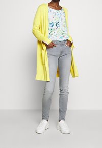 Mujer vestida con un cárdigan largo amarillo, blusa floral, jeans ajustados gris claro y zapatillas blancas, de pie con las manos en los bolsillos.