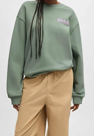 Personne portant un sweat-shirt vert sauge avec le logo "HUGO" en violet et un pantalon beige à taille élastique, avec de longues tresses sur l'épaule.