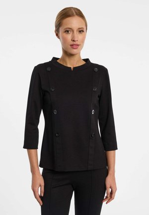 Woman with blonde hair wearing a black top with buttons along the front and three-quarter length sleeves standing against a white background.