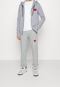 Sudadera gris con cremallera y un parche rojo, combinada con pantalones cargo gris claro que cuentan con bolsillos y un parche rojo similar. Camiseta blanca y zapatillas deportivas.