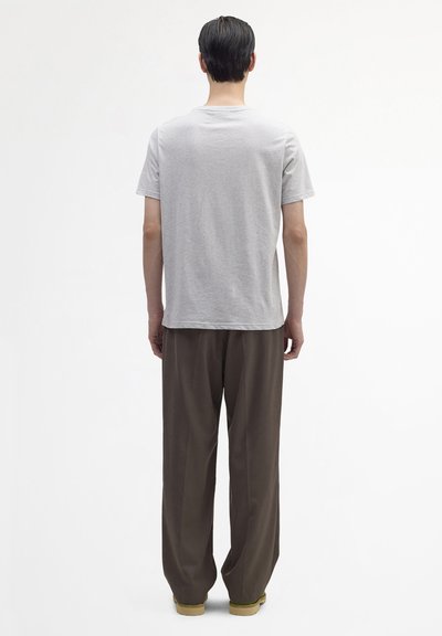 Homme debout, dos tourné, portant un t-shirt à manches courtes gris clair, un pantalon marron ample et des chaussures couleur sable, sur un fond blanc uni.