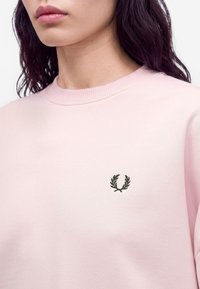 Femme portant un sweat-shirt rose pâle à col rond avec un petit logo de couronne de laurier vert foncé sur la poitrine gauche.