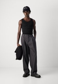 Young man wearing black tank top, gray wide-leg pants, black cap, holding a black jacket, standing against a plain light background.