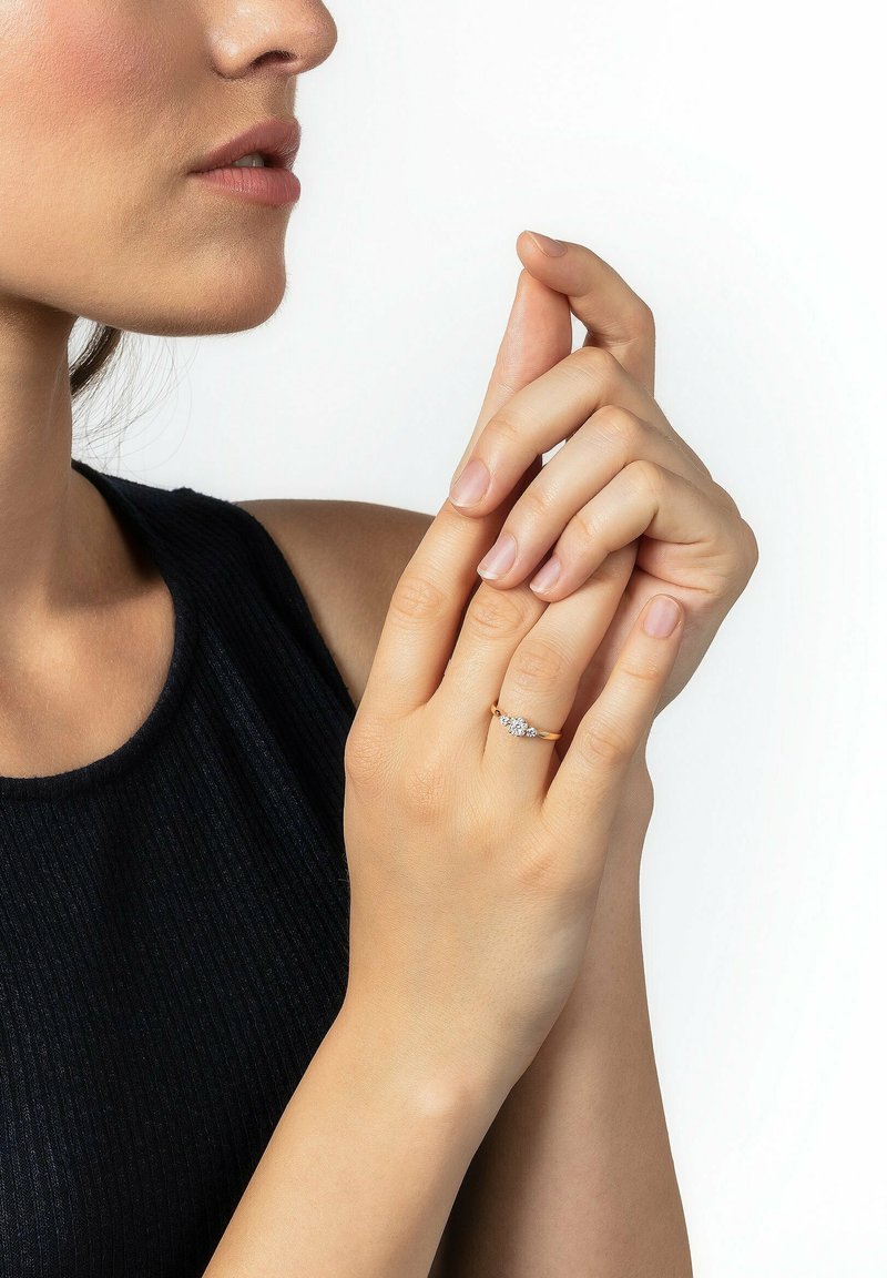 Goldring mit drei klaren Edelsteinen, getragen an einer Hand. Der Hintergrund ist weiß und hebt das Design des Rings und die Fingerspitzen hervor. Die glatte Hauttextur ist sichtbar.