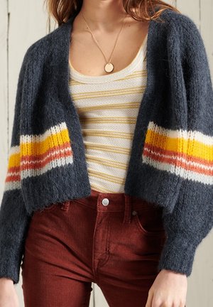 Femme portant un cardigan pelucheux bleu foncé avec des rayures jaunes, orange et blanches, une chemise texturée rayée blanche et jaune, et un pantalon couleur rouille.