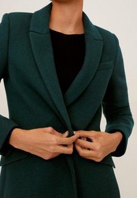 Blazer teal con colletto a punta, tessuto strutturato, tasca singola sul lato sinistro e vestibilità sartoriale, chiuso al centro.