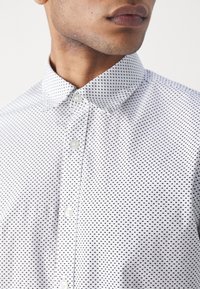 Camicia bianca con bottoni, caratterizzata da un piccolo motivo a quadri neri, colletto a punta e tessuto liscio. Leggera texture con bottoni chiari.