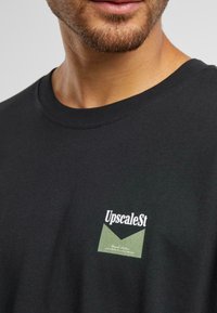 Black cotton t-shirt with a round neckline, featuring a small green and white logo patch on the left chest area. Smooth texture.