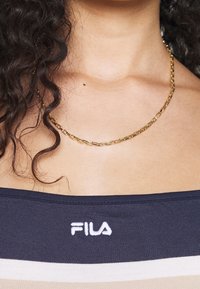 Złoty łańcuszek z wydłużonymi ogniwami. Na tle ciemnej, paskowanej bluzki z białym logo FILA. Gładka tekstura i błyszczące wykończenie.