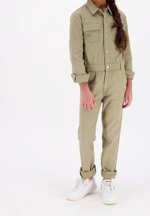 Combinaison vert olive clair avec deux poches poitrine, fermeture à boutons et taille élastique. Portée avec des baskets blanches. Texture de tissu lisse.