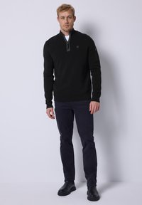 Suéter negro con cuello de cremallera y acabado texturizado, llevado sobre una camisa blanca, combinado con pantalones azul marino oscuro y botas negras de tobillo.