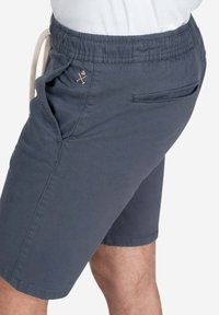 Pantalones cortos grises con cintura elástica, cordón, bolsillos laterales y un bolsillo trasero. Detalle sutil del logo en marrón claro en la parte delantera.