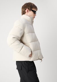 Geacă bej căptușită cu guler înalt și textură ribbed, având un fermoar frontal și buzunare laterale, asortată cu pantaloni negri.