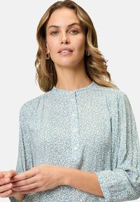 Helles blaues Blusenshirt mit einem strukturierten Muster aus kleinen, abstrakten Formen. Mit einem knöpfbaren Ausschnitt und langen Ärmeln mit Raffung.