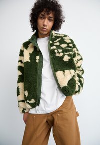 Casaco de fleece verde e bege com padrão de camuflagem, fecho de correr, gola alta e tecido texturizado. Usado sobre uma camisa branca e calças castanhas.