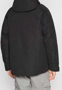 Svart isolerad jacka med huva, med en slät yta och elastiska ärmmuddar. Bakifrån visar jackan en avslappnad passform och minimala design detaljer.