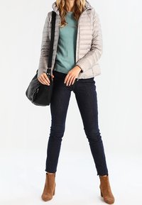 Jasnoszara kurtka puchowa z kapturem, turkusowy sweter, ciemne dżinsy i brązowe botki za kostkę, trzymająca czarną torebkę o teksturowanym wykończeniu.