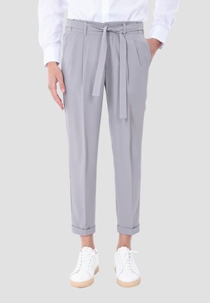 Persona che indossa pantaloni grigio chiaro con cintura annodata, orli risvoltati, scarpe da ginnastica bianche e camicia bianca a maniche lunghe.