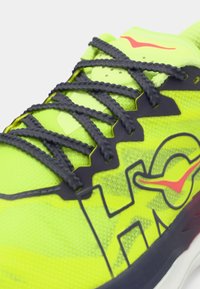 Εγγύς λήψη αθλητικού παπουτσιού νέον κίτρινο με μαύρα κορδόνια και κόκκινο λογότυπο σούφλας στη γλώσσα και το πλαϊνό πάνελ.